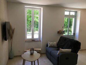 A living room with a sofa, a small table, a vase, and two windows with a view of greenery.