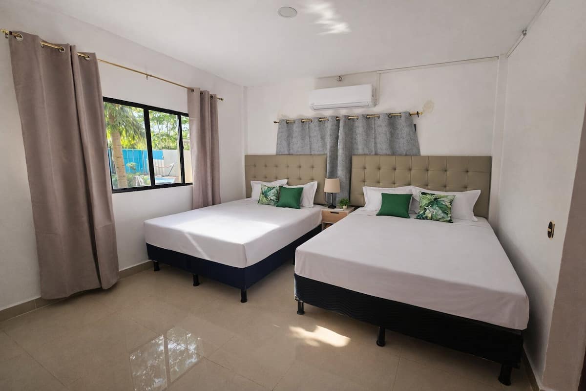 A hotel room with two beds, a window with curtains, an air conditioning unit, and a tiled floor.