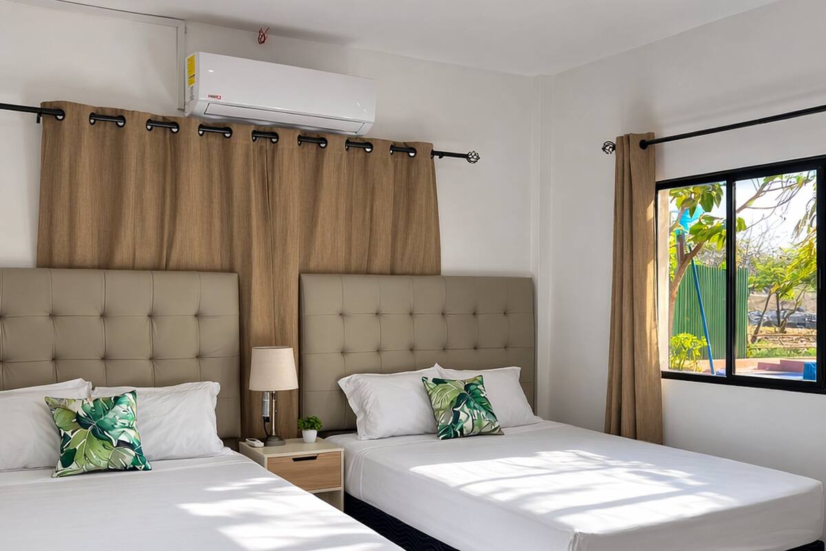 A hotel room with two beds, a window with curtains, an air conditioning unit, and a bedside table with a lamp.