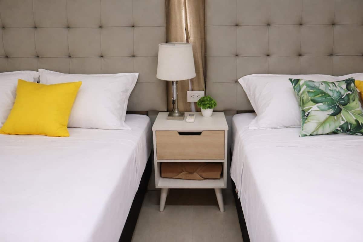 A hotel room with two beds, a nightstand with a lamp, a small plant, and a drawer.
