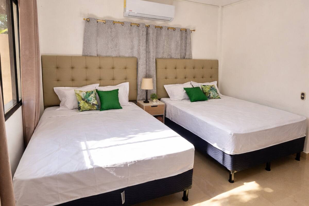 A hotel room with two beds, a window with curtains, an air conditioning unit, and a nightstand with a lamp.