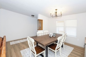 A dining room with a wooden table, white chairs, a chandelier, and a window with blinds.