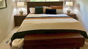 Quarto com cama, duas mesas de cabeceira com abajures, janela com persianas e vista para o verde lá fora.