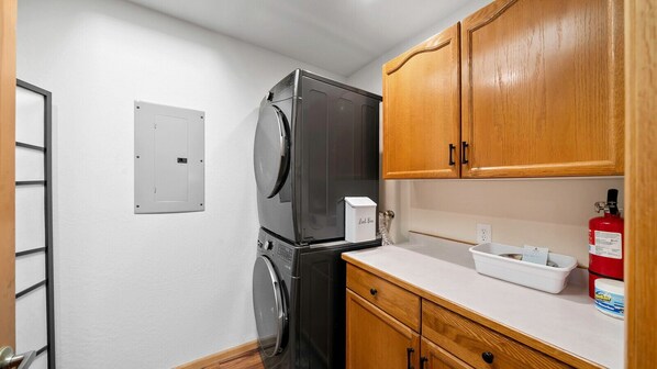 A compact kitchen with a refrigerator, a washing machine, wooden cabinets, a white countertop, and a fire extinguisher. - One36 Graeagle Meadows | Beautifully Updated, Mill Pond & Mountain Views (Graeagle)