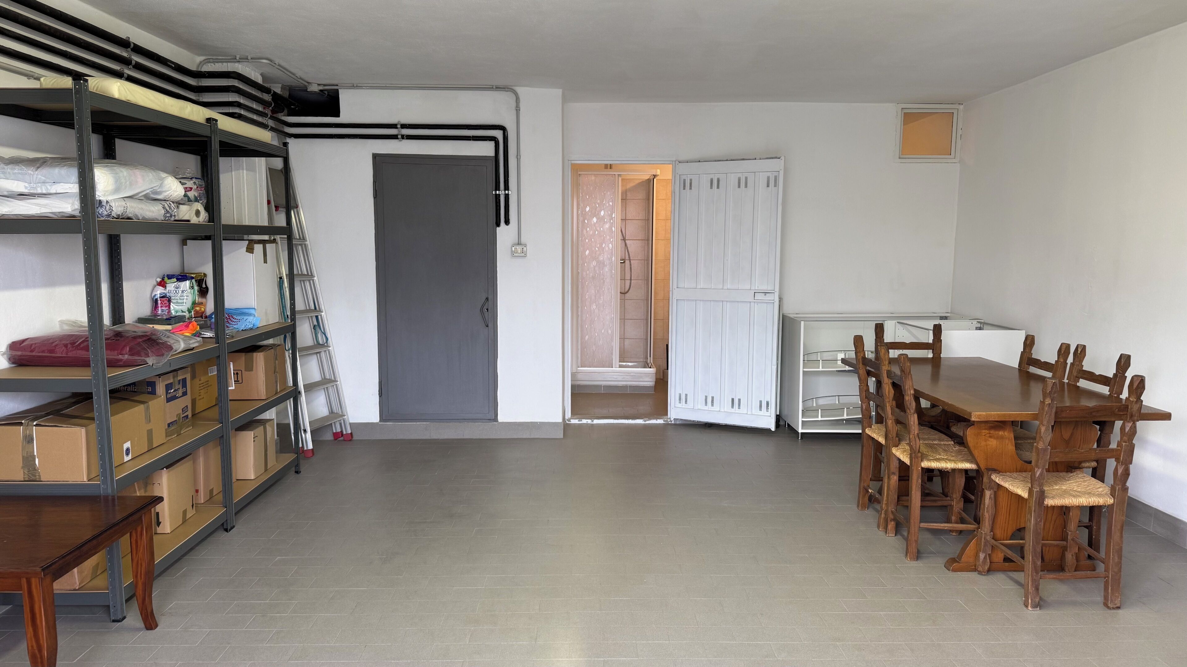 A room with a wooden dining table, chairs, a metal shelving unit, and a door leading to a shower area.
