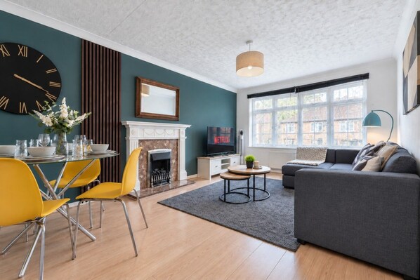 A modern living room with a fireplace, a dining area with yellow chairs, and a large wall clock. - Charming Flat • Great Location • Free Parking • Fast WiFi • Self-Check-In (Nottingham)