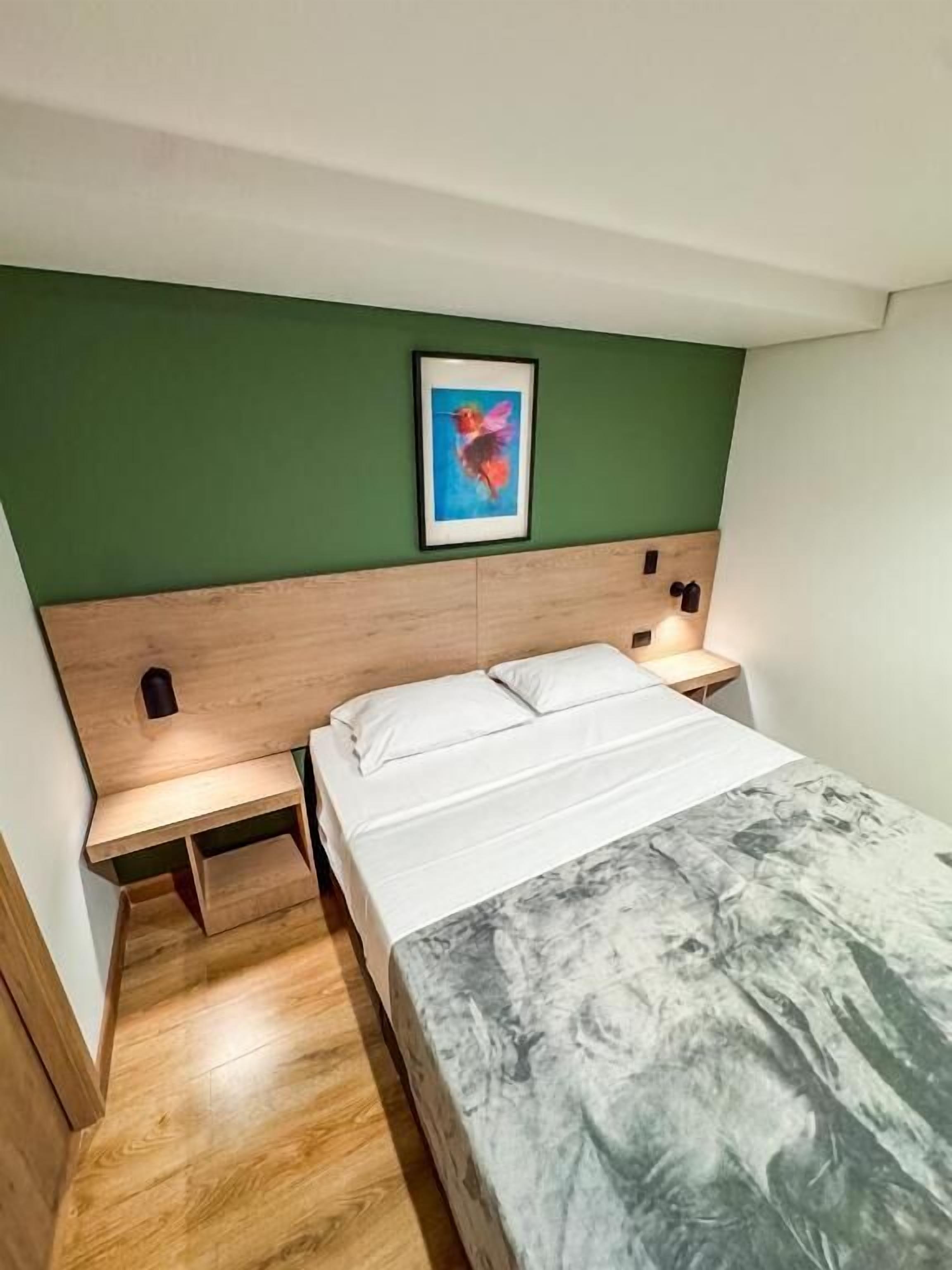 A bedroom with a wooden headboard, a bed with white bedding, a green wall, and a framed picture above the bed.