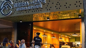 Ein Außenbereich von Tierra-Café mit Tischen und Stühlen, eine Person steht in der Nähe des Eingangs, und ein Gebäude mit einer gemusterten Fassade.