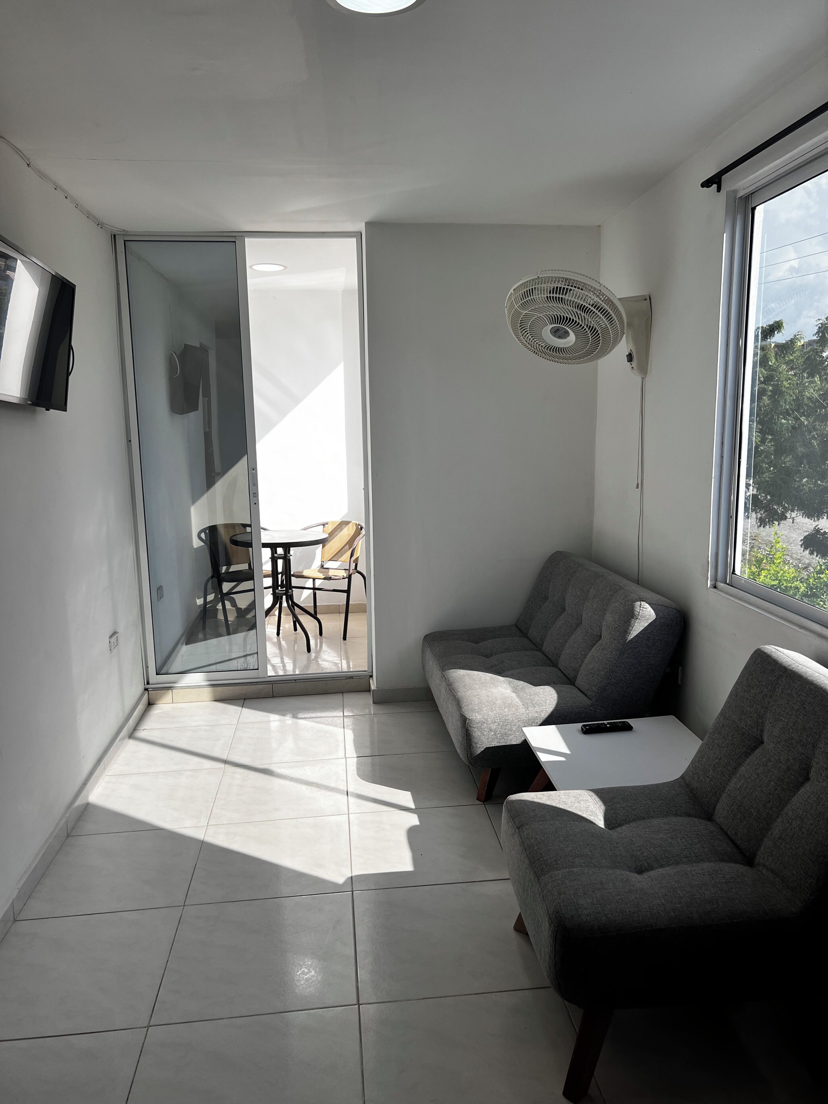 Et moderne værelse med en grå sofa, et lille bord, et fjernsyn ophængt på væggen og en skydedør, der fører til et udendørs område med et bord og stole.