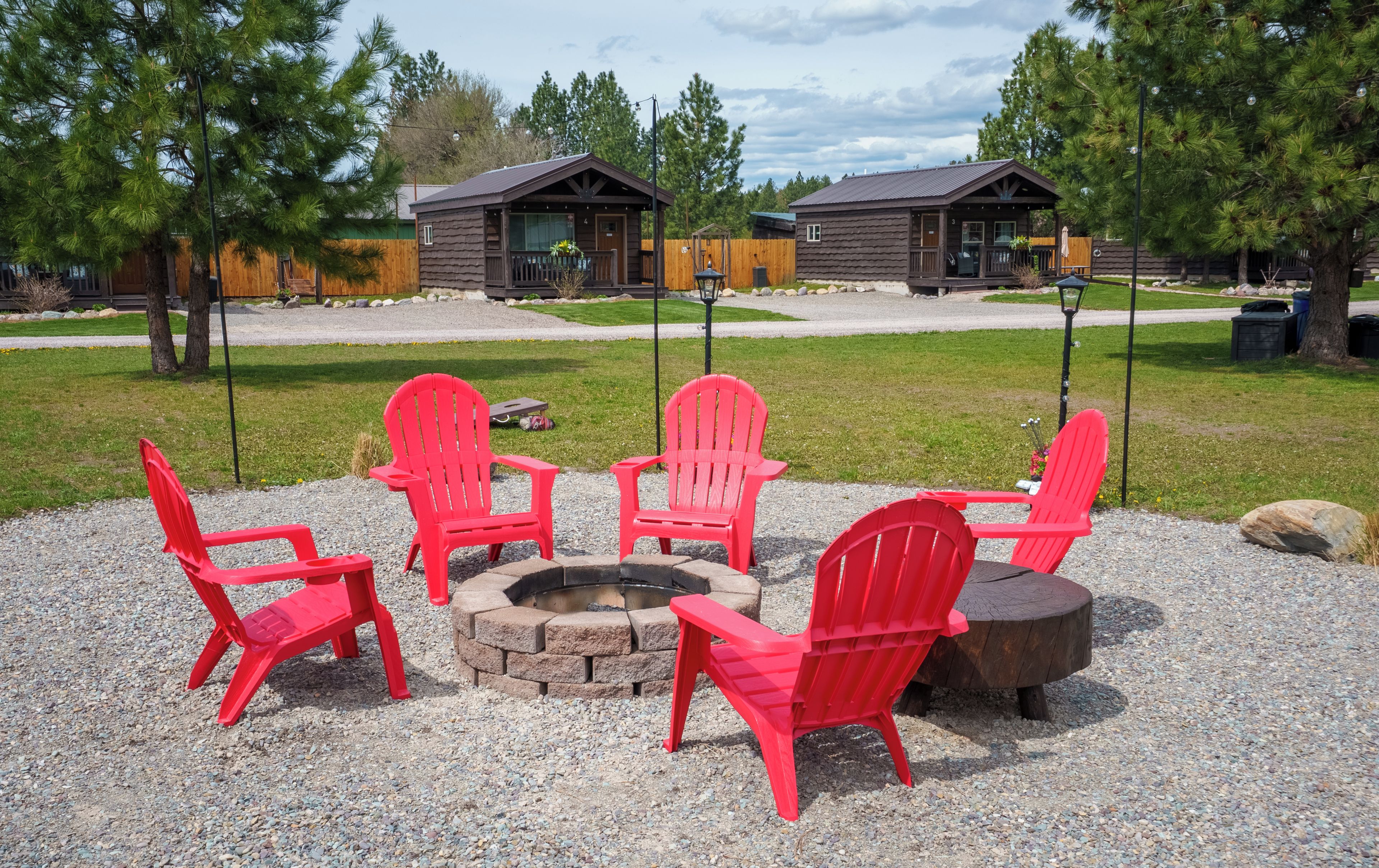 A fire pit surrounded by red Adirondack chairs in a gravel area with wooden cabins in the background.