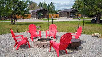 A fire pit surrounded by red Adirondack chairs in a gravel area with wooden cabins in the background.