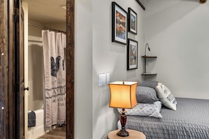 Quarto com cama, luminária de cabeceira, mesa de apoio, prateleira na parede e cortina de chuveiro.