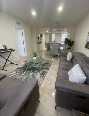 A modern living room with a sofa, a glass coffee table, and a dining area in the background.