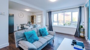 A modern living room with a sofa, a dining area, and a kitchen visible in the background. - Spacious 2 Bedroom House | Sleeps 8 | Driveway Parking | Pool Table & Games (Greater Manchester)