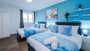 A modern hotel room with two beds, a large window, a blue accent wall, and a cityscape painting. - Spacious 2 Bedroom House | Sleeps 8 | Driveway Parking | Pool Table & Games (Greater Manchester)