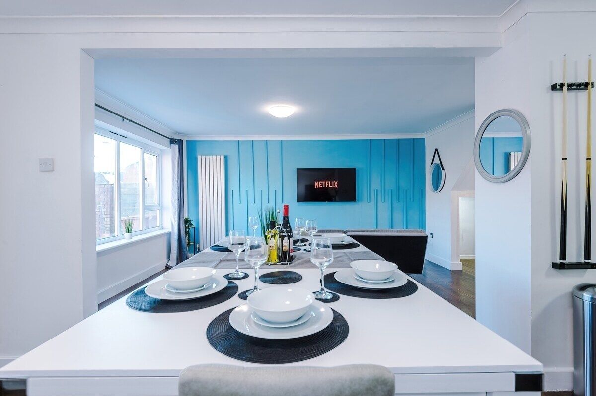 Un espace repas moderne avec une longue table dressée pour six personnes, un téléviseur fixé au mur et une fenêtre laissant entrer la lumière naturelle.