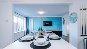 A modern dining area with a long table set for six, a television mounted on the wall, and a window providing natural light. - Spacious 2 Bedroom House | Sleeps 8 | Driveway Parking | Pool Table & Games (Greater Manchester)