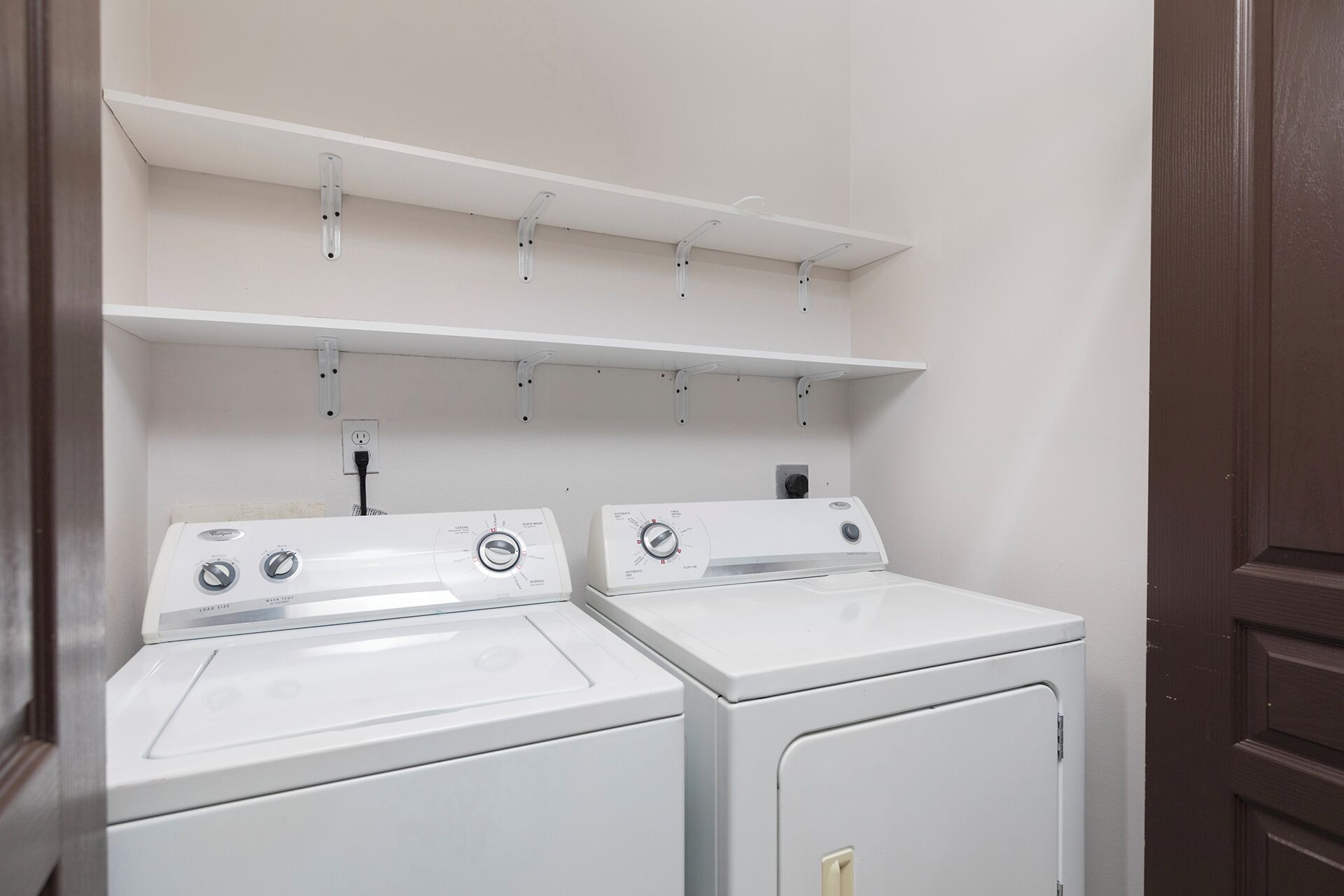 A laundry area with two white washers and dryers, a white shelf above, and a wooden door to the right.