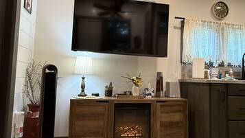 A living room with a flat-screen TV mounted on the wall, a fireplace, wooden flooring, and a kitchen area with a clock and window.