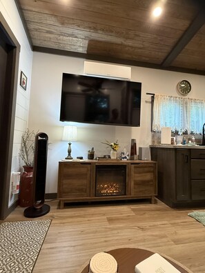 Sala de estar con televisor de pantalla plana montado en la pared, chimenea, piso de madera y una zona de cocina con un reloj y ventana.