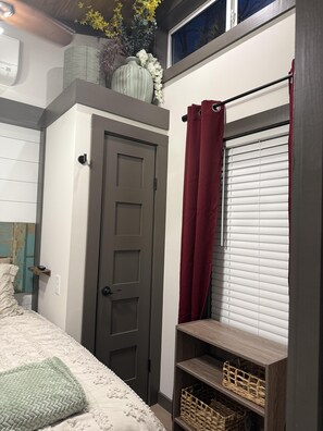 Un dormitorio con cama, una puerta, ventana con persianas, estante con cestas y un florero con flores en la pared.
