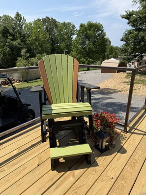 Una terraza de madera con una silla Adirondack verde, una mesita y una maceta con flores rojas.