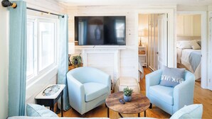 Ein Wohnzimmer mit zwei hellblauen Sesseln, einem runden Couchtisch, einem flachbildfernseher über einem Kamin und einem Bett im angrenzenden Zimmer.