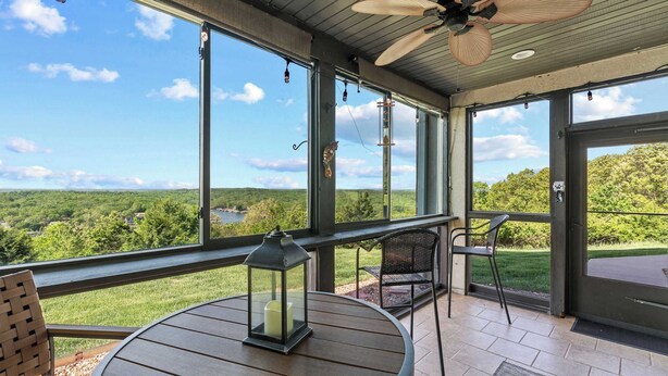 A covered patio with a table and chairs, large windows offering a view of a green landscape. - Sunset Ridge  Private Patio, Cliffside Lake Views (Lake Ozark)