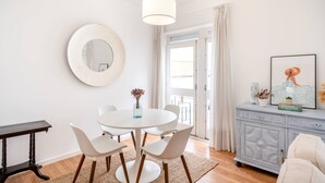 Un coin repas avec une table ronde, des chaises blanches, une table d’appoint en bois, une lampe blanche, un miroir, un meuble contenant des objets décoratifs et une porte donnant sur l’extérieur.