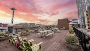 Una terraza en la azotea con piso de madera, sillas verdes, mesas de picnic, macetas con plantas y vistas de la Space Needle y el paisaje urbano.
