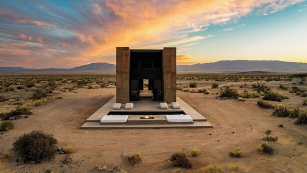 A desert landscape with a modern structure featuring a central seating area and a fire pit. - Folly Mojave Off-Grid Wilderness Retreat (Twentynine Palms)