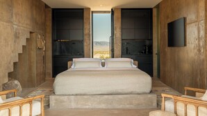 A bedroom with a large bed, a view through an open door, and a TV mounted on the wall. - Folly Mojave Off-Grid Wilderness Retreat (Twentynine Palms)