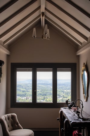 A room with a large window, a chair, a table with decorative items, and a ceiling with wooden beams and a hanging light fixture.