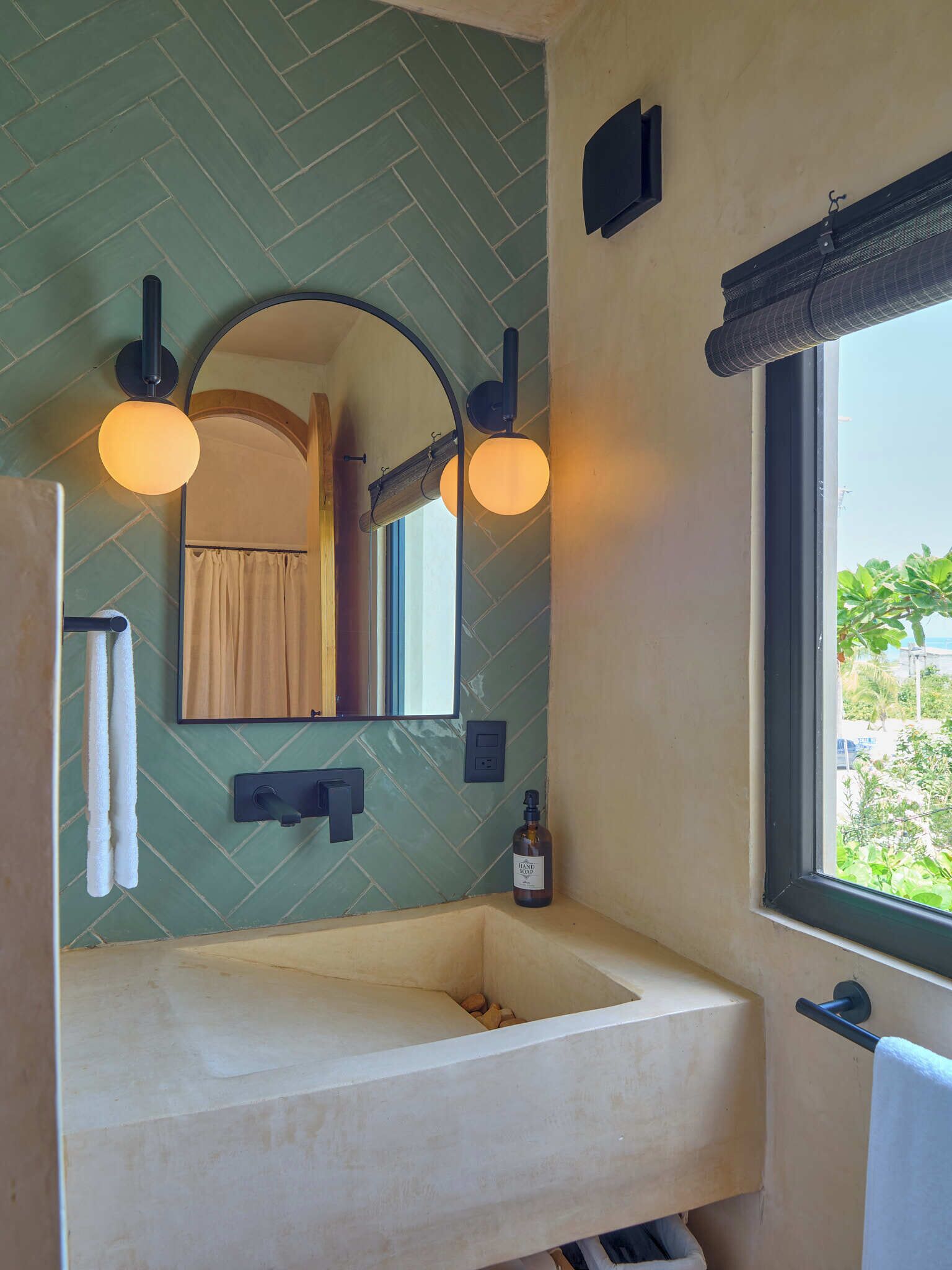 A bathroom with a white sink, two wall-mounted lights, a mirror, and a window with a view of greenery.