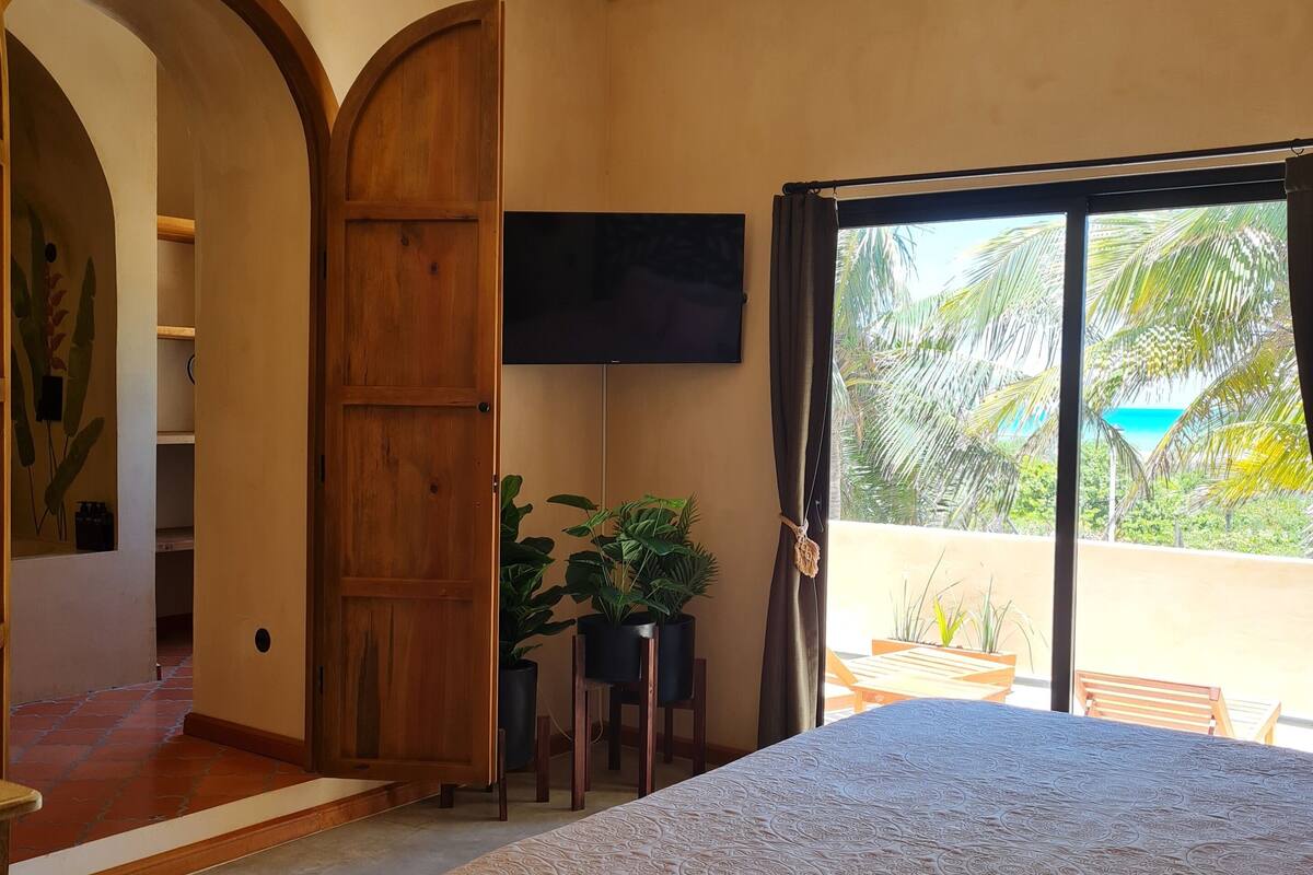 A bedroom with a bed, a TV, a balcony with plants, and a wooden door.