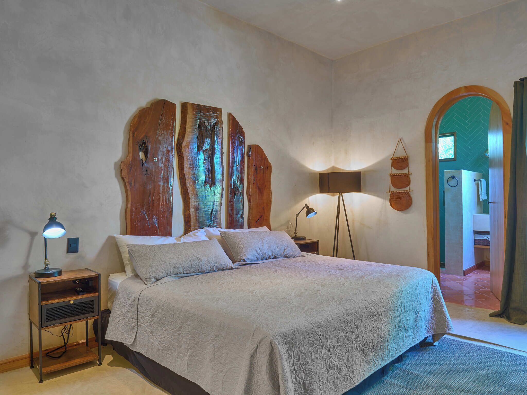 A bedroom with a large bed, two bedside lamps, a wooden headboard, and a view of a bathroom through an archway.