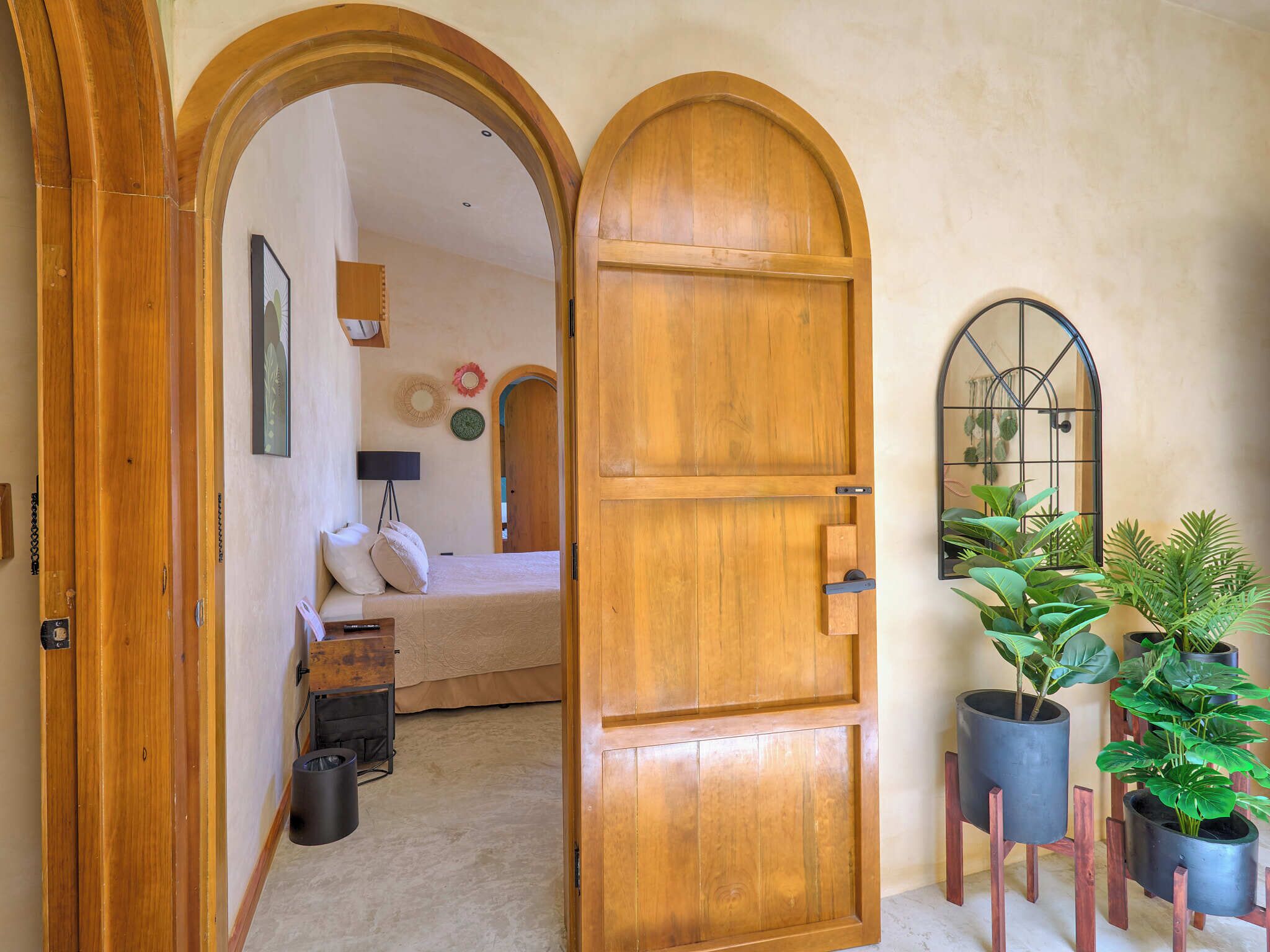 A room with a bed, a desk, and a window with plants.