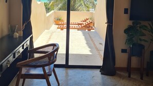 Habitación con puerta de cristal corredera que da a un balcón con vistas a palmeras y al océano. Hay una silla de madera y una mesita con una lámpara.