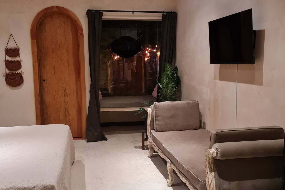 A bedroom with a bed, a sofa, a TV, a wooden door, and a window with curtains.