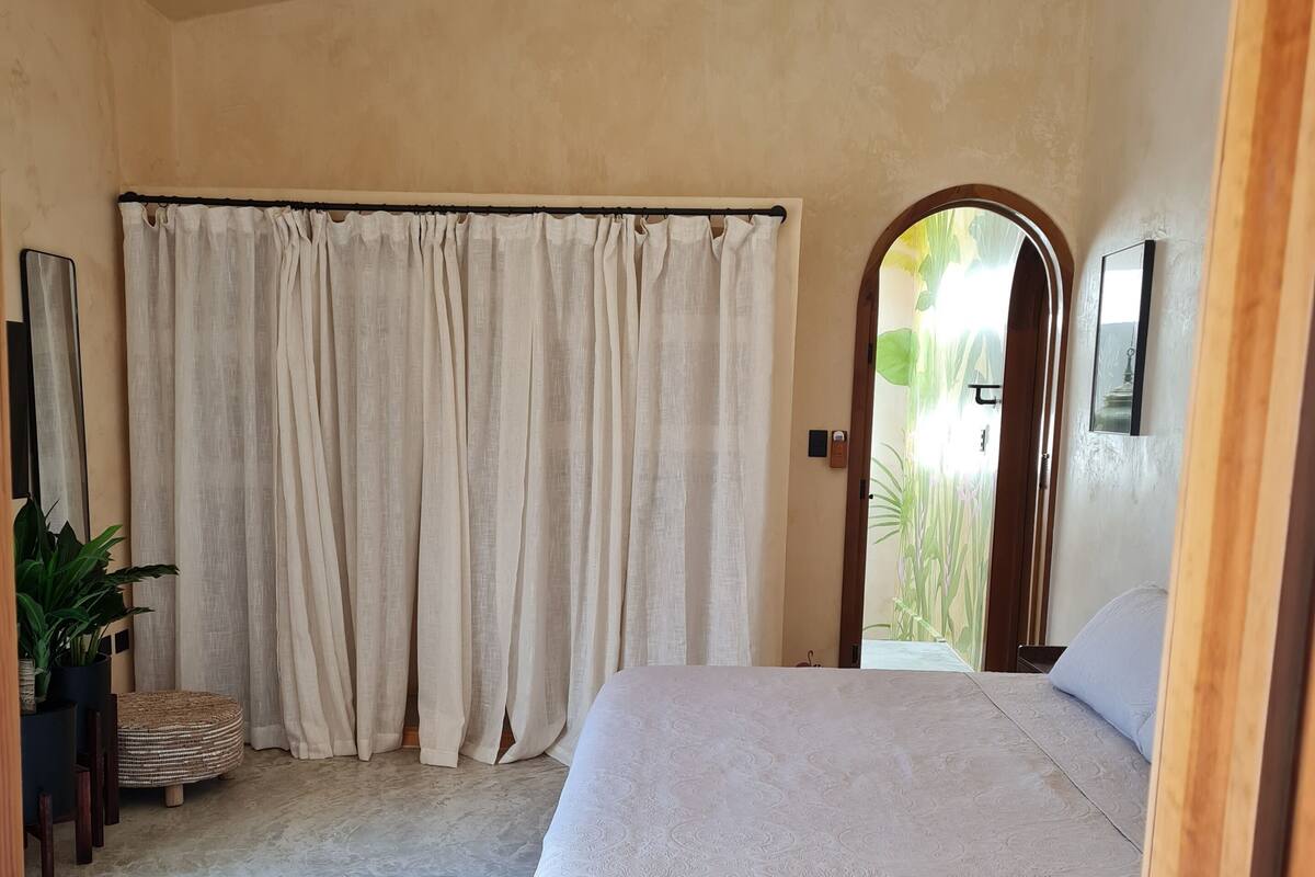 A bedroom with a bed, curtains, a mirror, a plant, and a door leading outside.