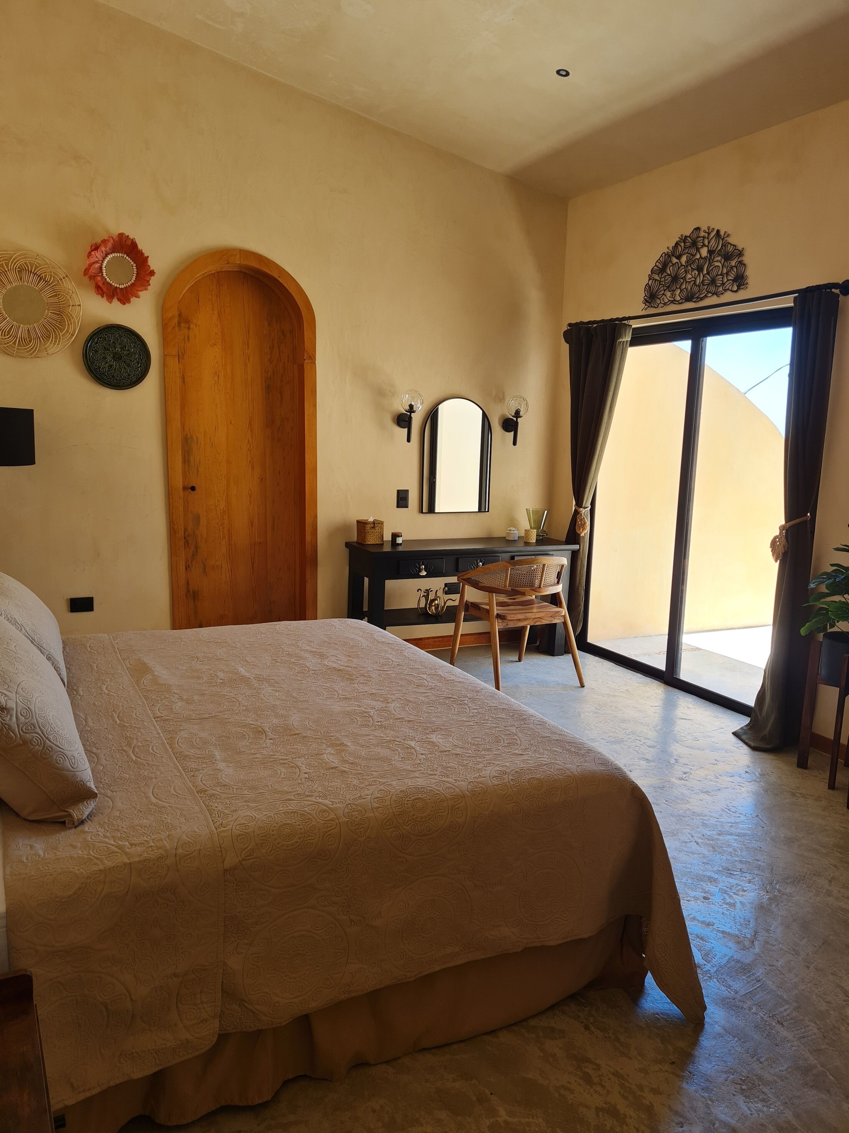 A bedroom with a bed, a wooden door, a desk with a mirror, and a view through a large window.