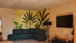 Une chambre à coucher avec un lit, un canapé vert, une télévision, une console et un mur orné d’une fresque aux motifs tropicaux.