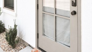 Ein weißes Gebäude mit Glastür und -fenstern, ein kleiner Garten mit Pflanzen und die Hausnummer 105 an der Wand.
