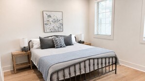 Ein Schlafzimmer mit einem Bett, zwei Nachttischen, einem Fenster und einem an der Wand hängenden Kunstwerk.