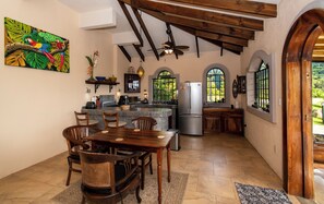 A kitchen with a dining area, wooden ceiling beams, a refrigerator, and a window with a view of greenery. - Stunning ocean and mountain views in Uvita! 1 bedroom house in the jungle (Uvita)