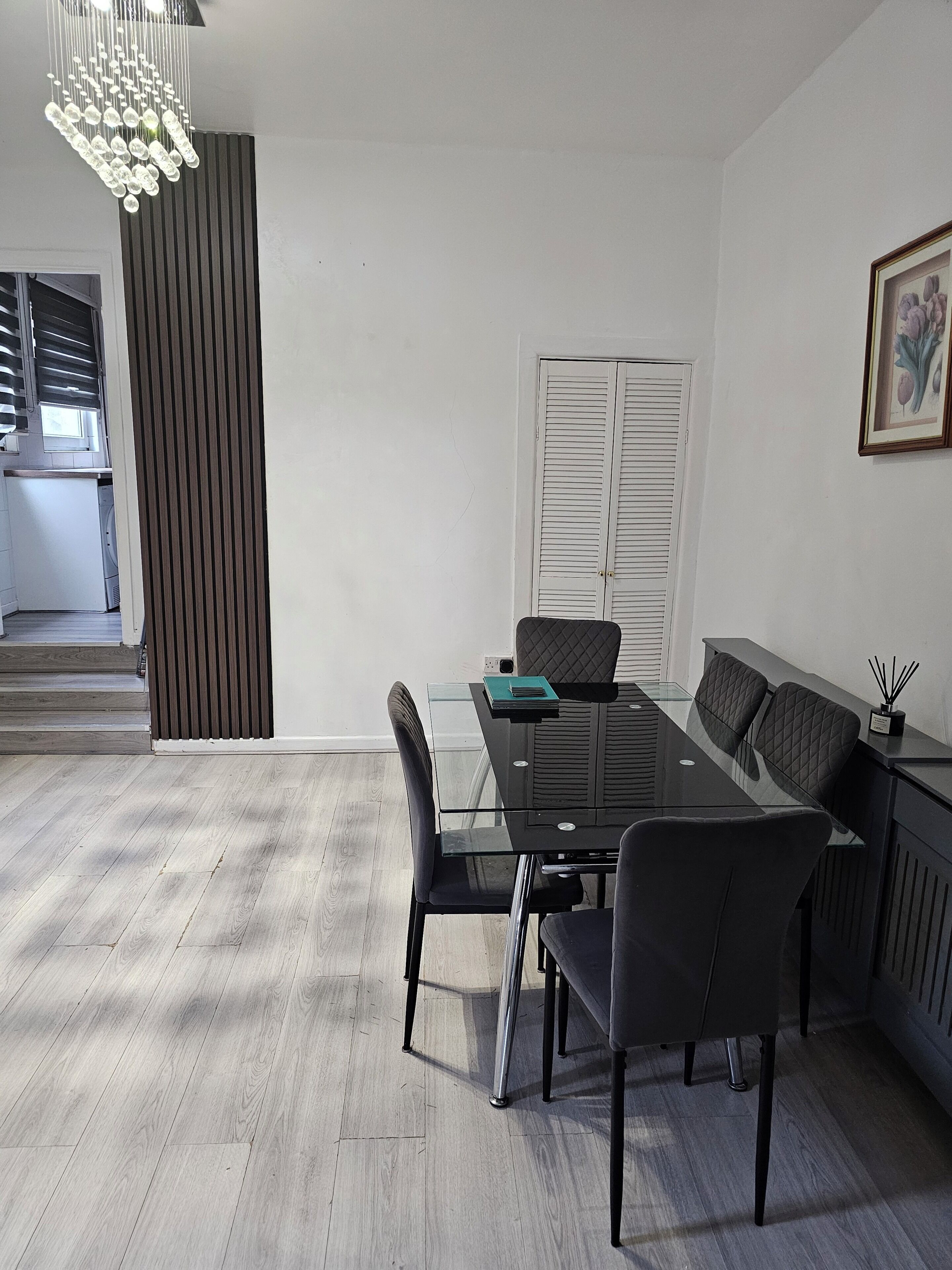 A modern dining area with a glass table, black chairs, a chandelier, and a view of a kitchen and a staircase.