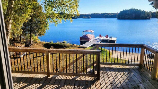 Eine Holzterrasse mit Geländer, die auf einen See blickt, eine Anlegestelle mit einem überdachten Boot und eine bewaldete Uferlinie.