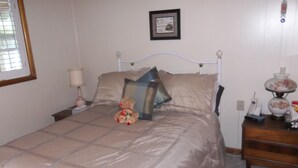 Ein Bett mit einer beigen Decke, ein Nachttisch mit einer Lampe, ein Teddybär und ein gerahmtes Bild an der Wand.
