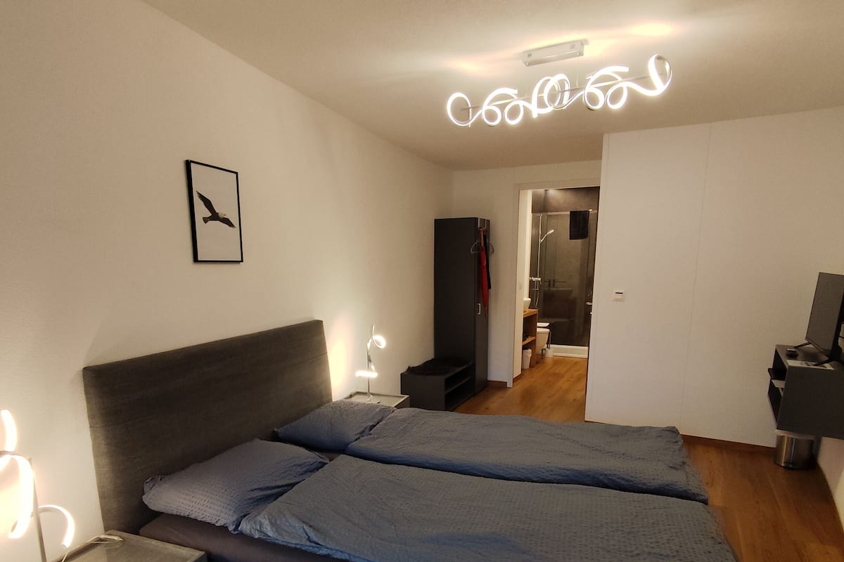A modern bedroom with a bed, bedside lamps, a framed picture, and a bathroom visible through an open door.
