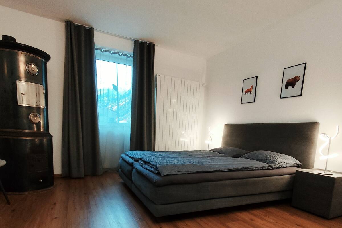 Un dormitorio con cama, cómoda, mesita de noche, ventana con cortinas y dos cuadros en la pared.